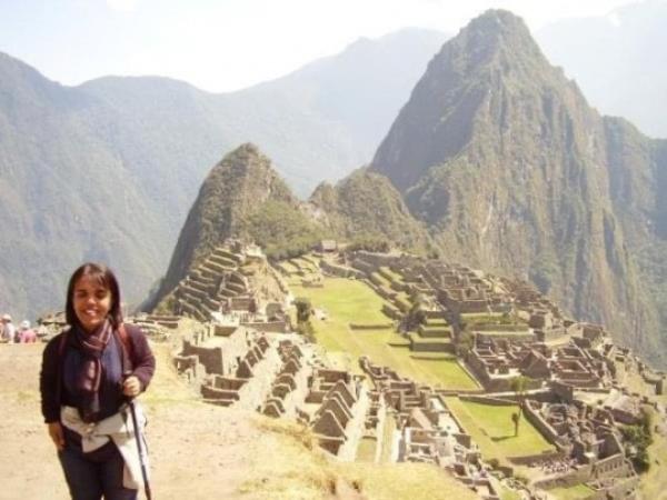 Roma on her hike, with hiking poles, wearing a scarf and dark clothes. Behind her is a landscape with ruins and mountains.